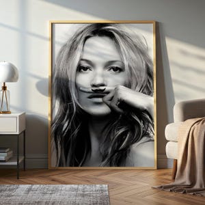 May include: A black and white portrait print in a thin wooden frame featuring a person with long hair holding a finger with a small black moustache graphic horizontally across their upper lip, creating a playful visual effect in this wall art.