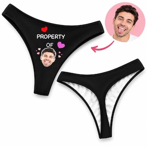 May include: Black thong underwear featuring the white text PROPERTY OF and a custom printed face of a man with his tongue out, surrounded by small red and pink heart graphics. The design is shown on the front and back of the garment.
