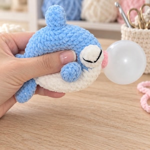 Könnte beinhalten: Ein gehäkeltes Plüsch-Delfin-Spielzeug aus hellblauem und weißem Garn. Der Delfin hat eine kleine, runde, weiße Blase am Mund, die eine Kaugummiblase nachahmt. Die Textur ist weich und flauschig, mit einem abgerundeten Körper und Flossen.