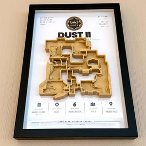 Könnte beinhalten: Ein gerahmtes 3D-Wandbild mit einer beigefarbenen Reliefkarte des Spiellevels Dust II. Der weiße Hintergrund enthält schwarzen Text mit Spieldaten wie Veröffentlichungsdatum, Entwickler, Kartentyp, Spieleranzahl und Standort, eingefasst in einem schwarzen Rahmen.