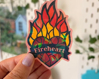 May include: A die-cut sticker featuring a heart shape topped with stylized flames. The design uses a stained-glass aesthetic with red, orange, yellow, and dark blue segments. A dark blue banner across the center displays the text Fireheart.