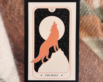 May include: A rectangular card featuring a black and cream illustration of a wolf howling at a full moon. The design includes the Roman numeral IV at the top and the text THE WOLF at the bottom, framed by thin orange lines on a black border.