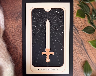 May include: A rectangular tarot card featuring a black background with a central illustration of a sword in peach and white. The card displays the Roman numeral II at the top and the text THE SWORD at the bottom, framed by thin lines.