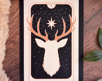 May include: A rectangular tarot card featuring a cream-colored silhouette of a stag head with large antlers against a black background. Above the stag is a white star with radiating lines. The text THE STAG is printed at the bottom with a Roman numeral I.