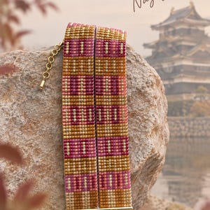 Op de afbeelding: Een brede kralenarmband met een geometrisch patroon in roze, goud en bruine tinten. De armband heeft een goudkleurige metalen staaf aan de onderkant met twee karabijnsluitingen en een verstelbare ketting. De tekst luidt KiraBanouDesigns Nagoya.