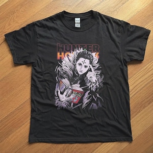 May include: A black short-sleeve t-shirt featuring a graphic print of a character with dark hair and a cross symbol on their forehead, holding an open book. The text HUNTER X HUNTER is printed in orange and purple above the character illustration.