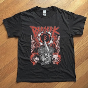 Op de afbeelding: Een zwart t-shirt met korte mouwen en een grafische print met de rode tekst BERSERK bovenaan. Het ontwerp bevat een centraal personage dat een groot zwaard vasthoudt, een cirkelvormig symbool en twee extra gezichten in rode en zwarte tinten.