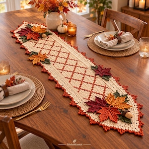 Pode incluir: Um caminho de mesa de crochê na cor creme com um padrão de losangos e borda festonada. O caminho é decorado com folhas de bordo de crochê tridimensionais em tons de vermelho, laranja e verde, juntamente com pequenas bolotas marrons.