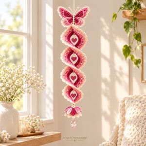 May include: A vertical hanging crochet decoration featuring a top butterfly, a spiral of five scalloped segments with heart cutouts in shades of pink and white, and a bottom flower with a dangling clear crystal bead. Text reads Design by Misbahkamal.