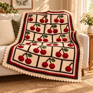 Puede incluir: Una manta de ganchillo rectangular con un patrón de cuadrícula repetitivo de cerezas rojas con hojas verdes sobre un fondo crema. La manta está enmarcada por un borde marrón oscuro y una franja roja gruesa, terminada con un borde de pompones crema.