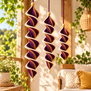 May include: Three hanging spiral ornaments made of crocheted yarn in deep purple and golden yellow, featuring a cream-colored border. Each piece is suspended by a purple cord with a wooden bead at the top and a small cream bead at the bottom.