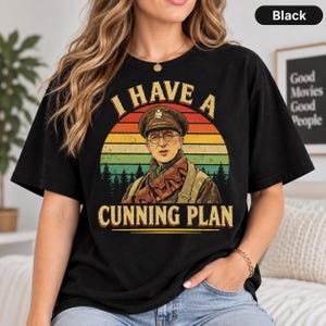 May include: A black short-sleeve t-shirt featuring a graphic print of a person in a military uniform and cap. The design includes a sunset background with pine trees and the text I HAVE A CUNNING PLAN in a bold, retro-style yellow font.