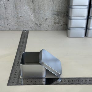 May include: A square silver metal tin with a removable lid, positioned next to a metal ruler showing a width of approximately 6.5 cm. The container has rounded corners and a smooth, matte finish, with a stack of identical tins visible nearby.