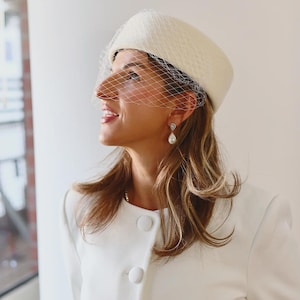 May include: A cream-colored pillbox hat featuring a delicate white birdcage veil that covers the upper face. The hat is paired with a matching cream-colored jacket that has three large, round, fabric-covered buttons down the front center.