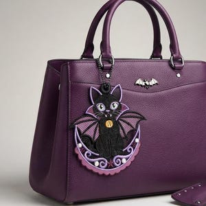 May include: A deep purple textured handbag featuring two top handles and a silver-toned metal bat emblem. A detachable embroidered charm depicts a black cat with bat wings sitting on a crescent moon, accented with small white beads and a star.