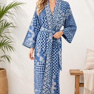 May include: A long-sleeved blue and white robe featuring a patchwork-style pattern of circles, dots, and botanical motifs. The garment has a V-neckline, wide sleeves, and a matching fabric tie belt at the waist, creating a wrap-style silhouette.
