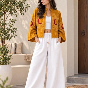 May include: A mustard yellow open-front jacket with flared sleeves and embroidered red crescent moon and sun ray motifs, paired with a white cropped tank top and white wide-leg trousers featuring a drawstring waist and side pockets.