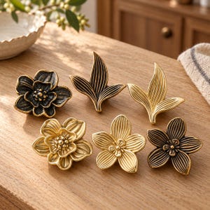 May include: A collection of six metal cabinet knobs featuring floral and leaf designs. The set includes three flowers with layered petals and two leaf-shaped knobs, finished in varying shades of antique gold and dark bronze metallic tones.