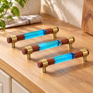 May include: Three cabinet pulls featuring a cylindrical design with a central section of translucent bright blue resin flanked by dark brown wood grain ends, all secured by brushed gold metal mounting posts at each end.