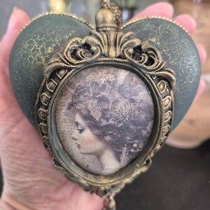Puede incluir: Un adorno en forma de corazón con una superficie texturizada de color verde oscuro y un marco dorado ornamentado. El centro presenta un retrato de perfil en tono sepia de una persona con cabello elaborado. Una pequeña cadena dorada y cuentas cuelgan debajo.