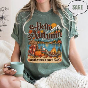 May include: A sage green t-shirt featuring a graphic print with the text Hello Autumn and Good Vibes & Cozy Days. The design depicts a wooden chair with a plaid blanket, pumpkins, a stack of books, a campfire, and a lantern by a lake.