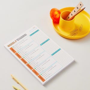 Può includere: Un blocco note Weekly Warrior con intestazioni arancioni e blu, dotato di sezioni per attività quotidiane, obiettivi e riflessioni. Un organizer da scrivania giallo contiene un righello in legno graduato fino a 15 cm, una matita e graffette.