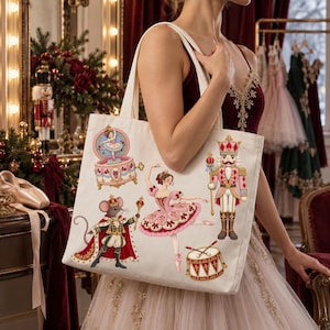 Puede incluir: Un bolso de tela color crema con un estampado colorido de ilustraciones temáticas del Cascanueces, que incluye una bailarina con tutú rosa, un rey ratón con capa roja, un soldado cascanueces, una caja de música y un tambor.