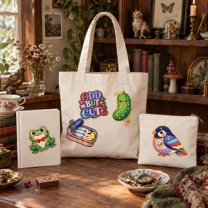 May include: A cream-colored canvas tote bag, a matching notebook, and a zippered pouch. The tote features embroidered patches of a pickle, a sardine tin, and the text "ODD BUT CUTE". The notebook shows a frog, and the pouch displays a colorful bird.