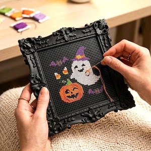May include: A square black ornate frame holds a black plastic grid with a cross-stitch design featuring a white ghost wearing a purple hat, an orange jack-o'-lantern, two candy corns, and purple bats, with a needle and orange thread.