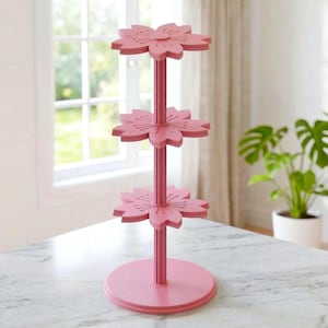 Puede incluir: Un expositor rosa de tres niveles con una columna central vertical y tres plataformas horizontales con forma de flor de cerezo. La estructura se apoya en una base circular, y cada nivel está separado por una sección de pilar estriado.