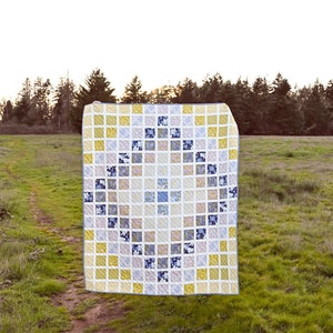 May include: A quilt with a geometric pattern in shades of yellow, blue, and gray. The quilt is made of squares and is hanging in a field.