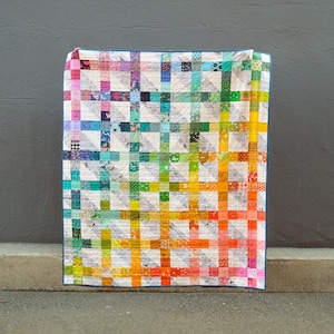 Może przedstawiać: Kolorowa kołdra patchworkowa z wzorem gradientowym tęczy. Kołdra jest wykonana z kwadratów z różnych tkanin w odcieniach różowego, fioletowego, niebieskiego, zielonego, żółtego i pomarańczowego. Kołdra jest złożona i leży na szarej betonowej powierzchni.