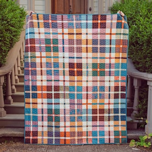 Könnte beinhalten: Eine farbenfrohe karierte Steppdecke mit blauem Rand. Die Steppdecke zeigt ein Patchwork-Design mit Quadraten in Blau-, Orange-, Braun-, Rosa-, Grün- und Weißtönen. Die Steppdecke wird im Freien vor einem Haus und Grünzeug präsentiert.