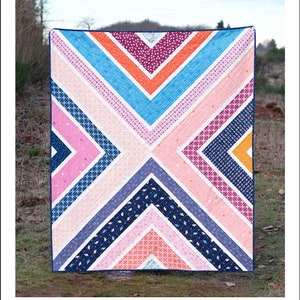 Op de afbeelding: Een kleurrijk quiltpatroon met een geometrisch ontwerp in tinten roze, blauw, oranje en wit. De quilt is gemaakt van een reeks diagonale strepen en wordt weergegeven op een grasachtige achtergrond. De tekst "The Sylvie Quilt" en "A PATTERN BY ERICA JACKMAN" staat bovenaan de afbeelding. De tekst "BABY, LAP, AND TWIN SIZES" staat onder de quilt. De tekst "KITCHEN TABLE QUILTING" staat onderaan de afbeelding.
