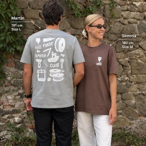 Könnte beinhalten: Ein hellgraues T-Shirt mit einer weißen Grafik auf der Rückseite, die 3D-Druck-Symbole und den Text FIRST LAYER CLUB zeigt, sowie ein braunes T-Shirt mit einem kleinen weißen 3D-Drucker-Düsen-Symbol auf der Brust, kombiniert mit schwarzer und weißer Hose.