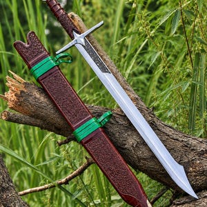 May include: A silver-colored steel sword with a pointed blade and a crossguard. The handle is brown with a circular pommel. The accompanying brown scabbard features intricate embossed patterns, two bright green bands, and a gold-toned metal tip.