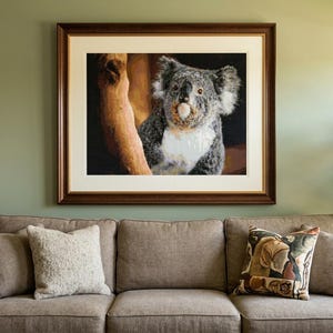 May include: A framed wall art print featuring a koala perched on a tree branch against a dark background. The artwork is enclosed in a dark brown wooden frame with a wide off-white matting, displayed above a beige sofa with several throw pillows.