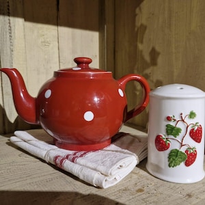 Op de afbeelding: Een glanzende rode keramische theepot met witte stippen en een bijpassend deksel, rustend op een gevouwen witte doek met dunne rode strepen, naast een witte cilindrische keramische strooier met een reliëfpatroon van aardbeienranken.