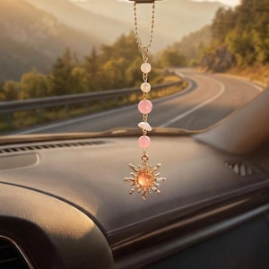 Op de afbeelding: Een auto-achteruitkijkspiegelhanger met een goudkleurige zon-bedel met een perzikkleurige steen in het midden. De bedel hangt aan een goudkleurige kogelketting met afwisselend ronde roze kralen en onregelmatige witte stenen als accenten.
