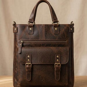 May include: A dark brown leather vertical tote bag featuring two top handles, a zippered horizontal pocket, and a front flap pocket secured with two brass-toned buckles. The initials D.L. are embossed on the front flap, and the bag includes side D-rings.