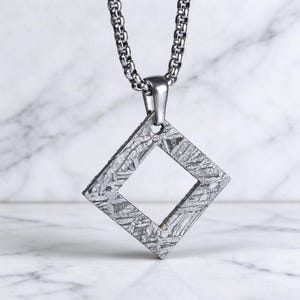 Op de afbeelding: Een vierkante hanger met een opvallend Widmanstätten-patroon, hangend aan een zilverkleurige metalen boxketting. De hanger heeft een hol midden en een metallic grijze afwerking met ingewikkelde, elkaar kruisende kristalstructuren.
