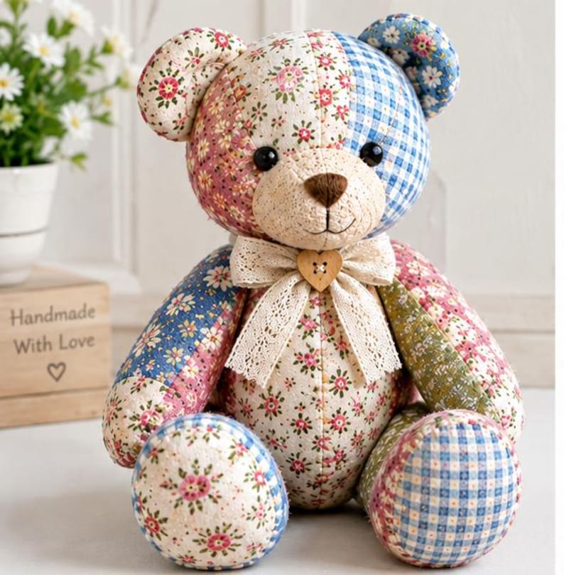 May include: A patchwork teddy bear constructed from various fabric patterns including pink floral, blue gingham, and blue floral prints. The bear features black button eyes, a brown stitched nose, and a cream lace bow with a heart-shaped wooden button.