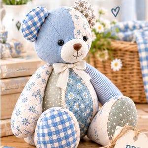 May include: A patchwork teddy bear crafted from various blue and white fabrics featuring floral, gingham, and striped patterns. The bear has a cream-colored muzzle, a brown nose, and a cream ribbon bow. A tag attached to the bear reads PDF Pattern.