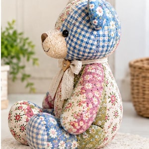 May include: A stuffed teddy bear crafted from various patchwork fabrics featuring blue gingham, floral patterns in pink, green, and white, and a cream-coloured muzzle. The bear wears a lace ribbon around its neck with a small wooden heart button.