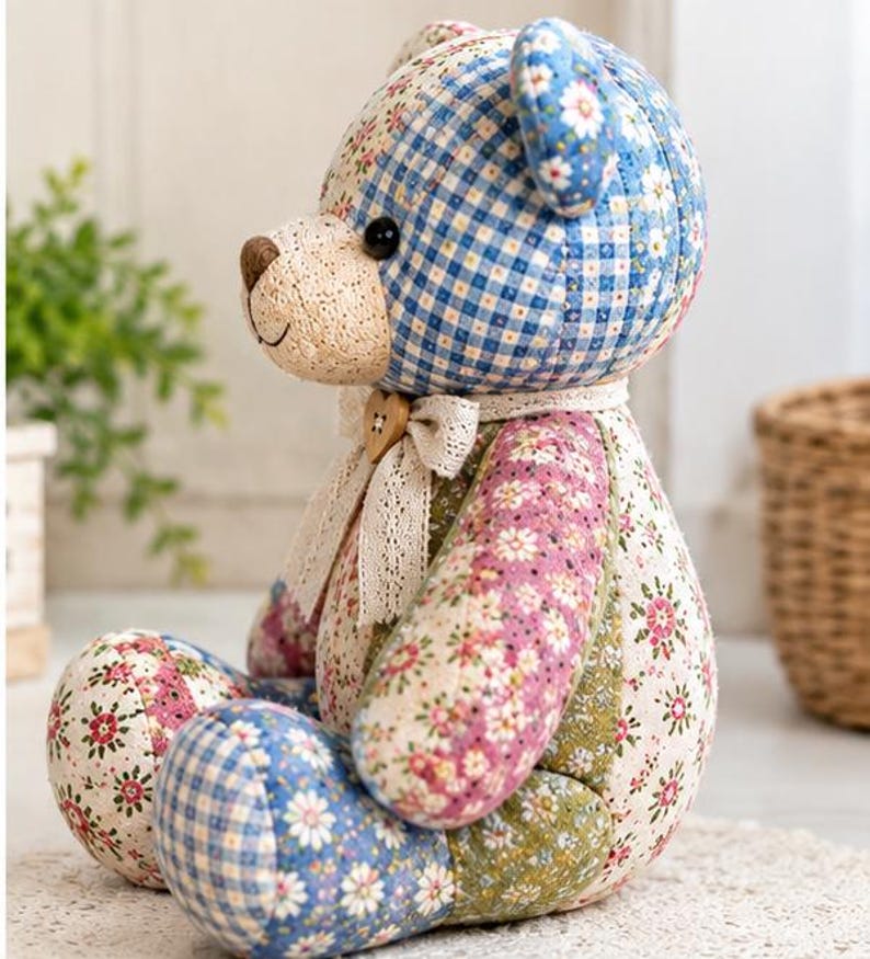 May include: A stuffed teddy bear crafted from various patchwork fabrics featuring blue gingham, floral patterns in pink, green, and white, and a cream-coloured muzzle. The bear wears a lace ribbon around its neck with a small wooden heart button.