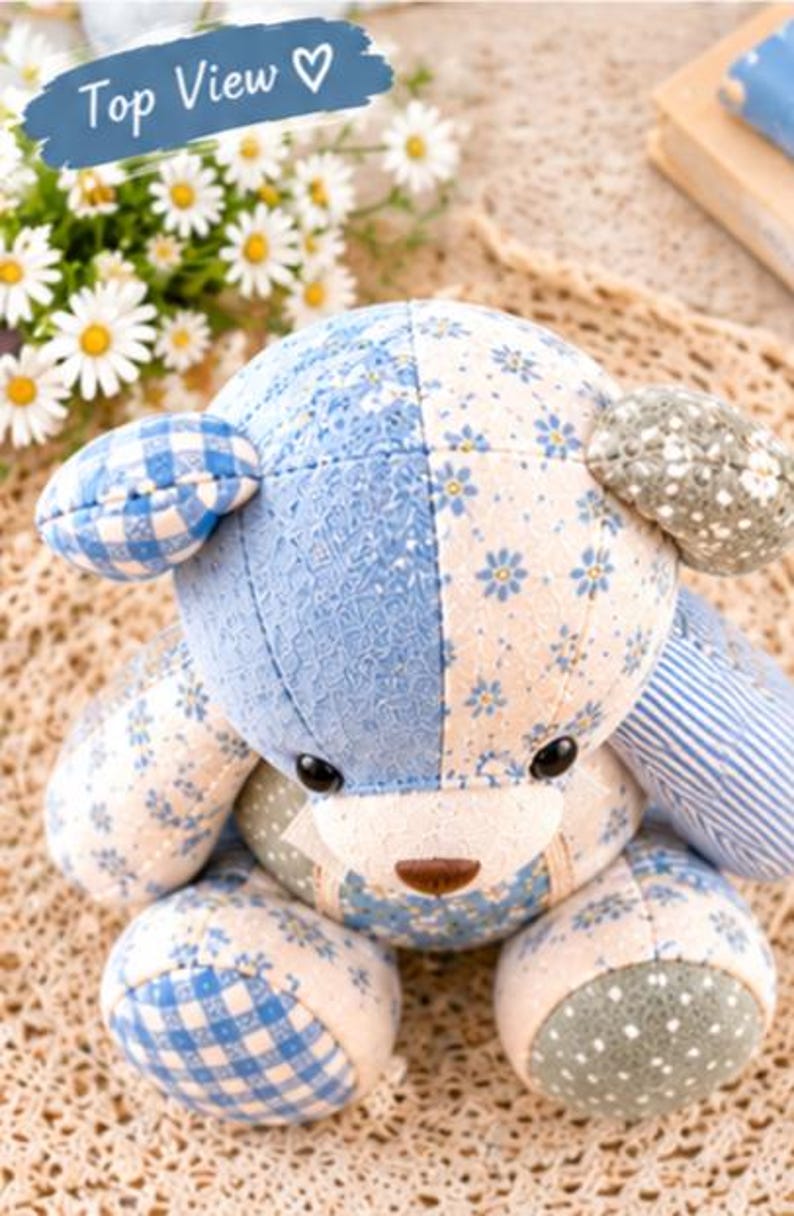 May include: A stuffed bear toy constructed from various fabric patches in shades of blue, white, and sage green. The bear features floral, gingham, and striped patterns, with black plastic eyes and a brown stitched nose, viewed from above.