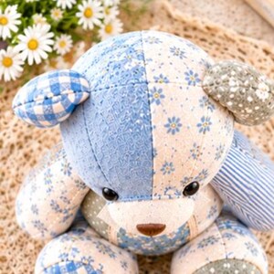 Peut inclure: Un ours en peluche compos&eacute; de divers morceaux de tissu dans des tons de bleu, blanc et vert sauge. L'ours pr&eacute;sente des motifs floraux, &agrave; carreaux et ray&eacute;s, avec des yeux en boutons noirs et un nez cousu marron, vu de dessus.