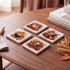 Puede incluir: Un juego de cuatro posavasos cuadrados blancos con ilustraciones de temática otoñal. Cada posavasos muestra un diseño único con calabazas, tazas de bebidas calientes, ramas de canela, piñas y follaje otoñal en tonos naranja y marrón.