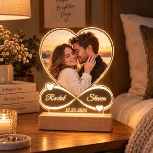 Może przedstawiać: Lampka nocna z akrylu w kształcie serca ze zdjęciem pary, symbolem nieskończoności, imionami Rachel i Steven oraz datą 25.12.2024. Lampka jest zamontowana na prostokątnej drewnianej podstawie i emituje ciepłe, delikatne światło.