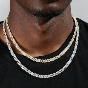 May include: Two layered cuban link chains, one in a gold tone and one in a silver tone, both featuring a pavé-set surface with small clear stones. The chains are worn around the neck, resting against a plain black fabric garment.